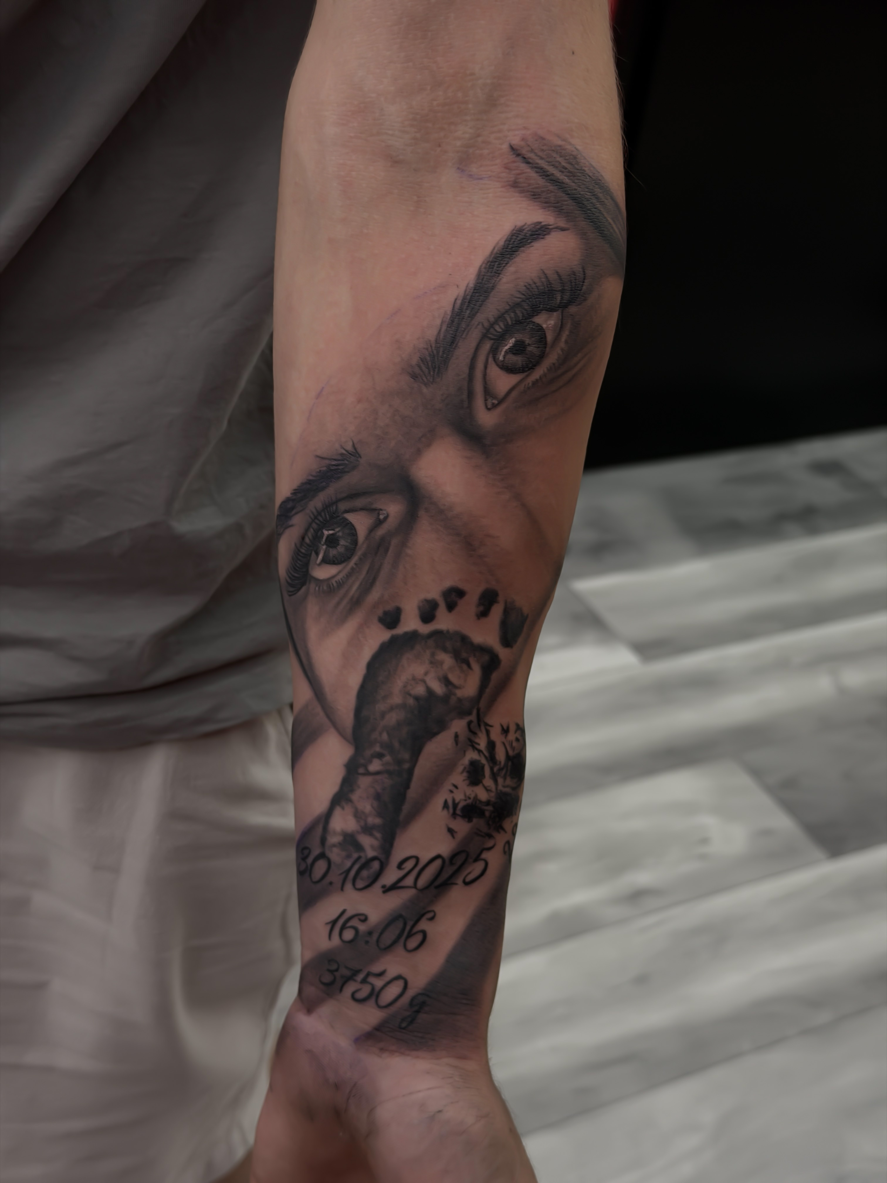 Tatuaj black & grey cu ochi și amprentă de picior pe antebraț